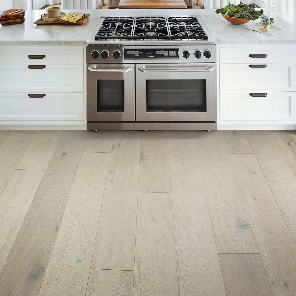 light grey white oak hardwood flooring in a  kitchen with a large white island, including a natural gas stove, white stone counters and dark brown hardware. On the counter is a bowl of kale and a wooden serving tray. 