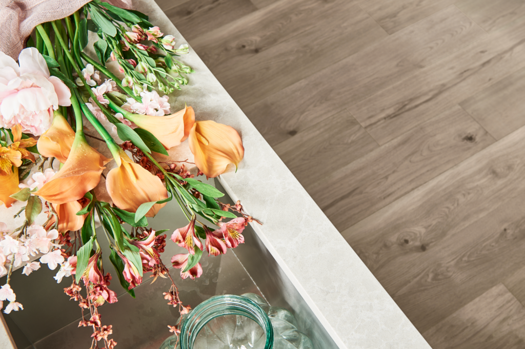 Looking down at beige Laminate flooring with a white table with orange and pink flowers on top.