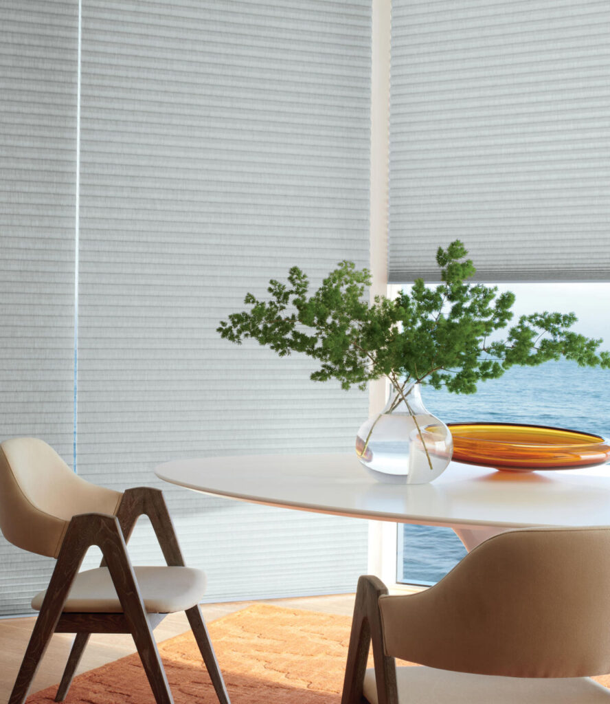 Honeycomb window blinds in a dining area, showing one blind open to let the light in and view of the ocean, and the other fully closed to protect the flooring. 
