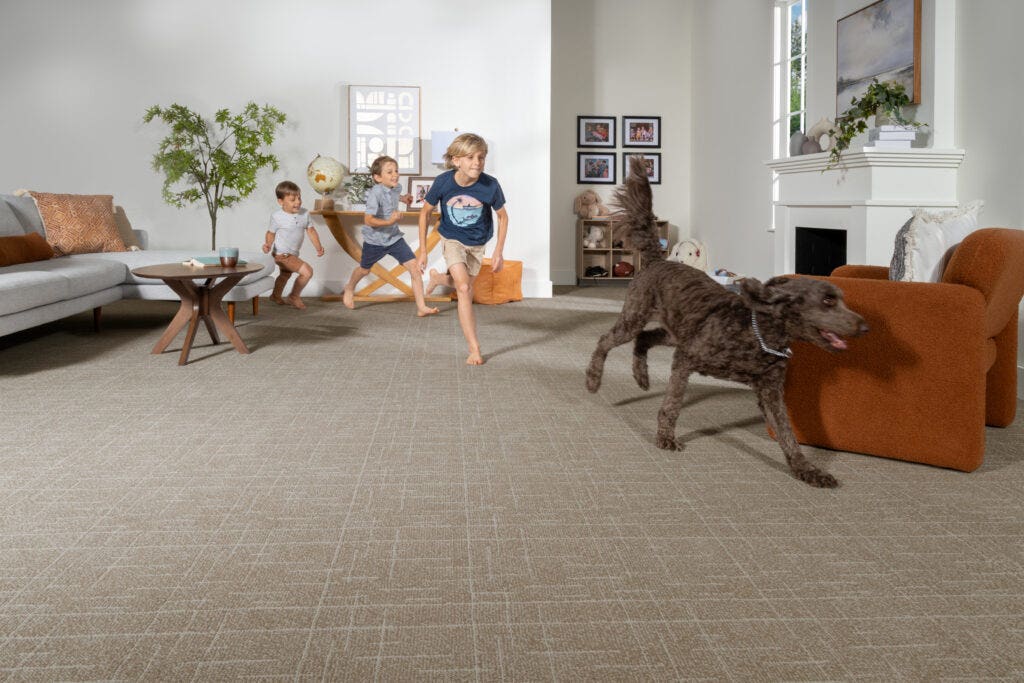 Kids and a dog run across a living room, complete with a couch, chair, and brown carpet.