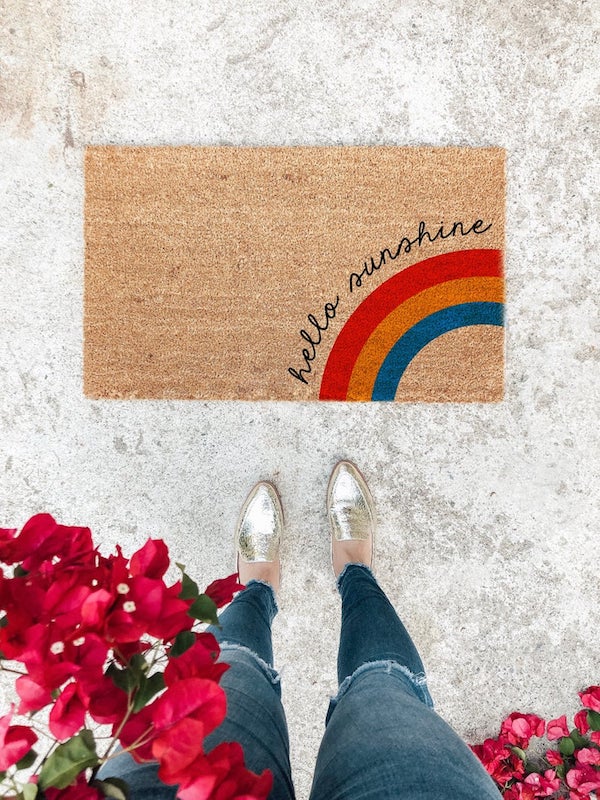 Looking down at Woman's feet that are beside a Doormat with a rainbow and the words "Hello Sunshine".