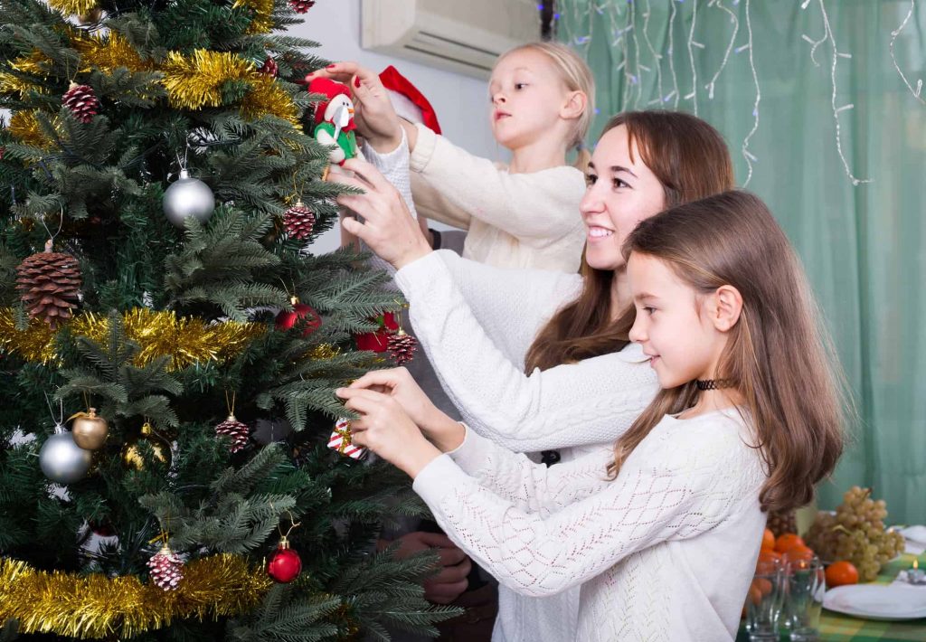 Cheerful united family with little daughters decorating Christmas tree together at home. Focus on girl