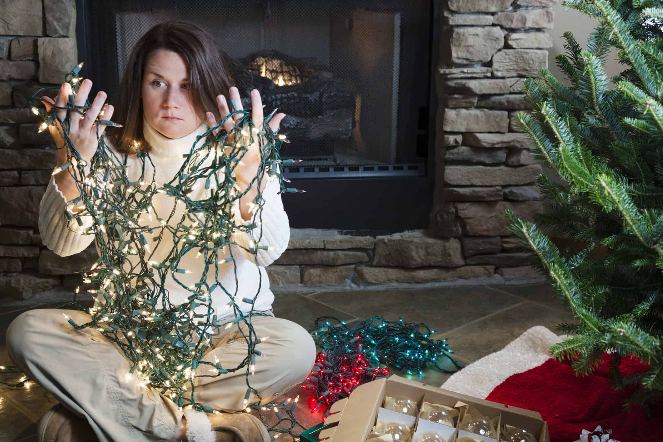 Young woman sitting next to Christmas tree holding entangled string of lights